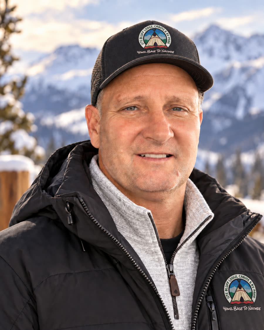 Johnny Shaw, owner of Ute Bluff Lodge, in a Ute Bluff cap and jacket in front of snowy peaks